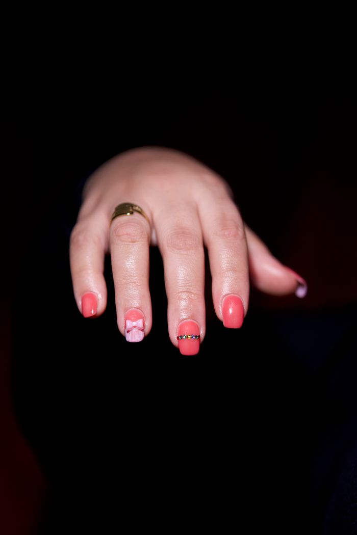 Close-up of a hand with intricate pink nail art and a gold ring against a dark background.